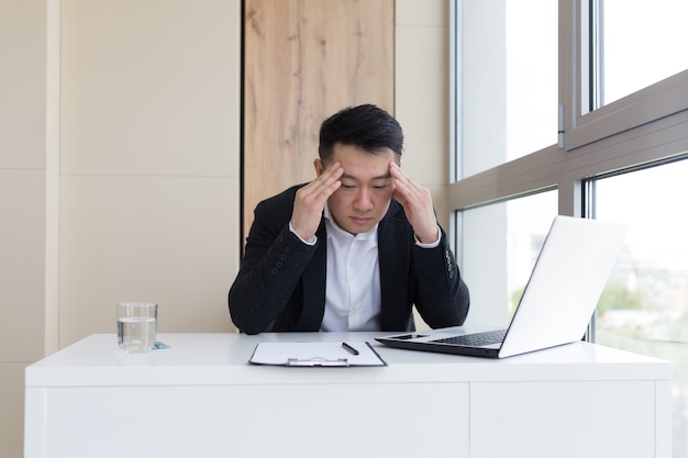 Junge asiatische Büroangestellte, die bei der Arbeit im Büro unter Kopfschmerzen leiden, trinkt Medizinpille mit Wasser. Kranker Mann im Anzug am Computer drinnen mit starken Schmerzen verwendet das Medikament am Arbeitsplatz