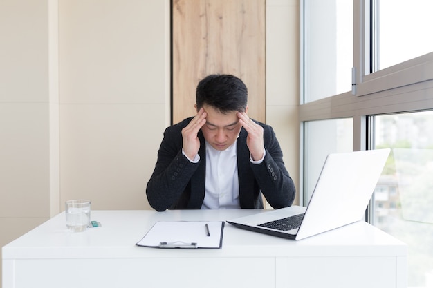 Junge asiatische büroangestellte, die bei der arbeit im büro unter kopfschmerzen leiden, trinkt medizinpille mit wasser. kranker mann im anzug am computer drinnen mit starken schmerzen verwendet das medikament am arbeitsplatz