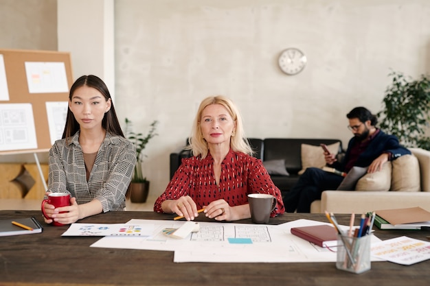 Junge asiatische Büroangestellte und ihre blonde reife Kollegin beim Kaffee beim Sitzen am Tisch mit vielen Papieren gegen Geschäftsmann