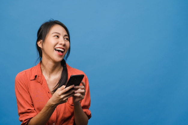 Junge asiatische Dame, die Telefon mit positivem Ausdruck benutzt, breit lächelt, in Freizeitkleidung gekleidet ist und sich glücklich fühlt und isoliert auf blauer Wand steht. Glückliche entzückende frohe Frau freut sich über Erfolg.