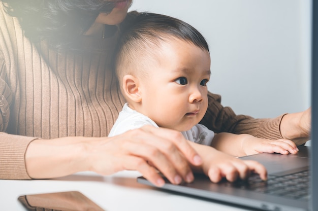 Foto junge asiatische frau arbeiten von zu hause aus, während babysitting spielen laptop im familienkonzept. mutter lehrt ihren sohn für die nutzung des internets.