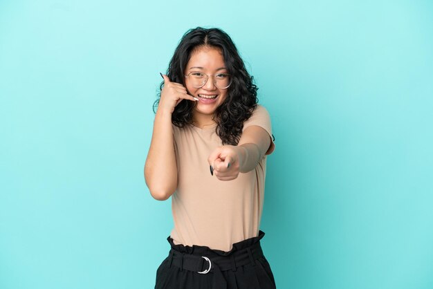 Junge asiatische Frau auf blauem Hintergrund isoliert, die Telefongeste macht und nach vorne zeigt