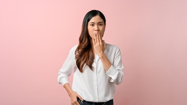 Junge asiatische Frau bedeckt ihren Mund mit ihrer Hand, die auf rosa isoliertem Hintergrund steht