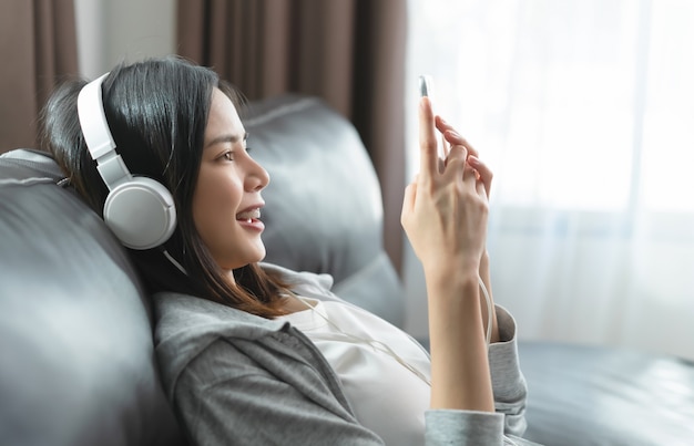 Junge asiatische Frau des glücklichen Lächelns, das Musik von weißen Kopfhörern hört und Smartphone zu Hause verwendet.