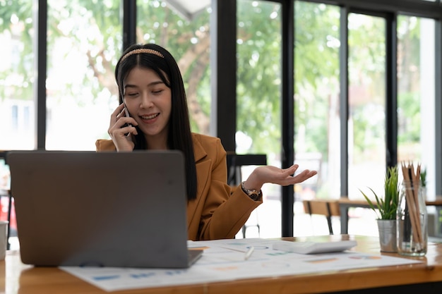 Junge asiatische frau, die am handy spricht und lächelt, während sie an ihrem arbeitsplatz im büro sitzt