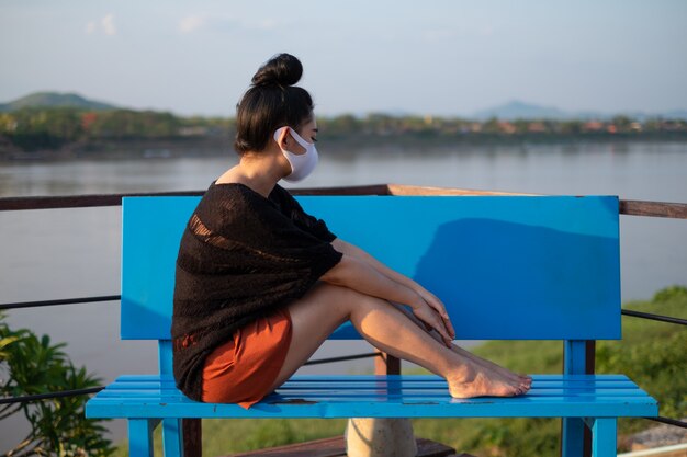 Junge asiatische Frau, die an der blauen Bank sitzt und eine medizinische Maske trägt, um vor Coronavirus zu schützen