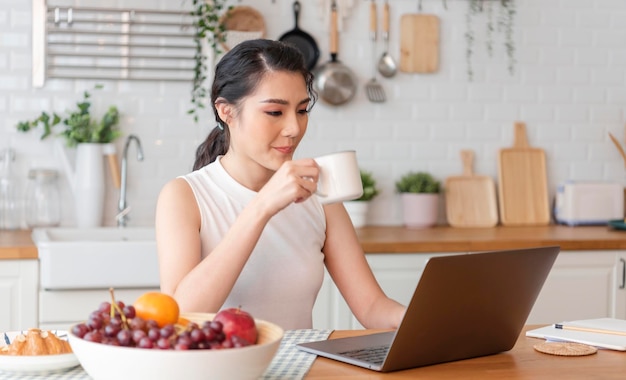 junge asiatische frau, die an einem laptop arbeitet, während sie im küchenraum sitzt und kaffee trinkt