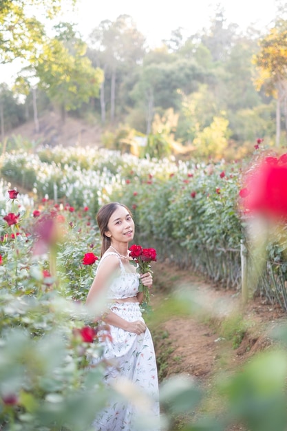 Junge asiatische Frau, die ein weißes Kleid trägt, posiert mit einer Rose im Rosengarten