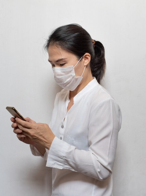 Junge asiatische Frau, die eine chirurgische Maske trägt, verhindern die Verwendung des auf weißem Wuhan-Coronavirus (COVID-19) isolierten Smartphones im öffentlichen Bereich. Gesundheitswesen und medizinisches Konzept. &