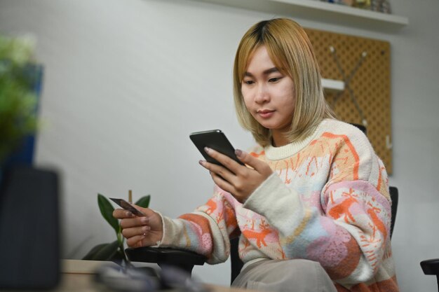 Junge asiatische frau, die eine kreditkarte hält und smartphone für online-einkäufe verwendet