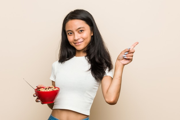 Junge asiatische Frau, die eine Müslischüssel lächelt und zeigt beiseite und zeigt etwas auf Leerstelle hält.