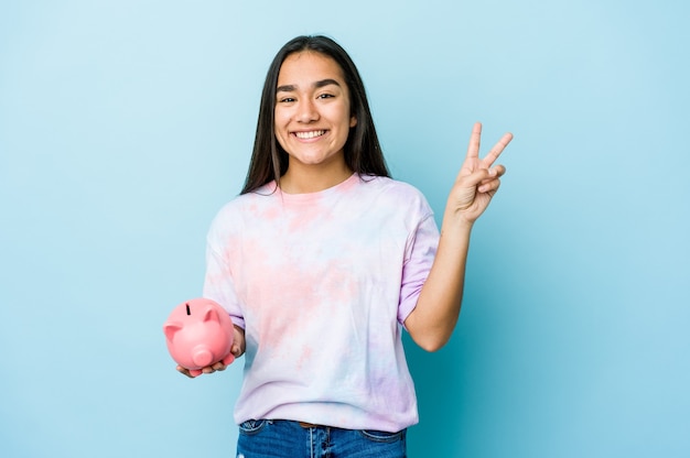 Junge asiatische Frau, die eine rosa Bank über der isolierten Wand freudig und sorglos hält und ein Friedenssymbol mit den Fingern zeigt.