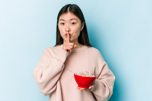 Junge asiatische Frau, die eine Schüssel Müsli isoliert auf blauem Hintergrund hält, ein Geheimnis bewahrt oder um Stille bittet