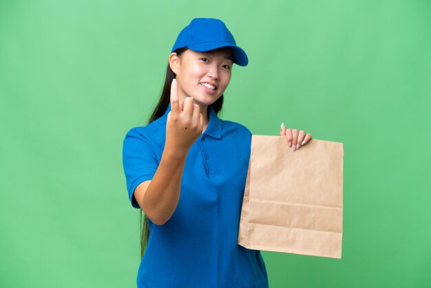 Junge asiatische Frau, die eine Tüte Essen zum Mitnehmen vor isoliertem Hintergrund nimmt und eine kommende Geste macht