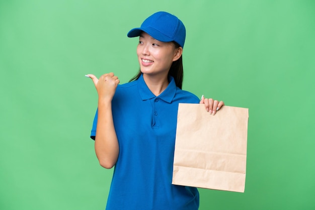 Junge asiatische Frau, die eine Tüte mit Essen zum Mitnehmen vor isoliertem Hintergrund nimmt und zur Seite zeigt, um ein Produkt zu präsentieren