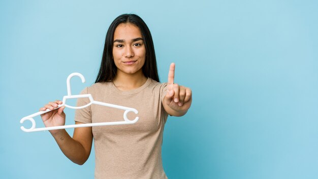 Junge asiatische Frau, die einen Aufhänger lokalisiert auf blauer Wand hält, die Nummer eins mit Finger zeigt.