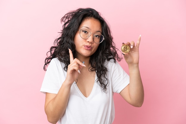 Junge asiatische Frau, die einen Bitcoin einzeln auf rosafarbenem Hintergrund hält und beabsichtigt, die Lösung zu realisieren, während sie einen Finger nach oben hebt