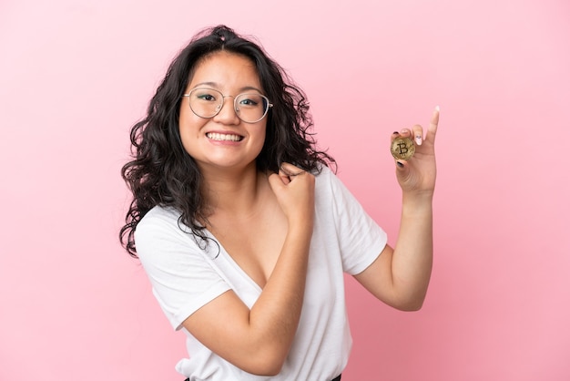 Junge asiatische Frau, die einen Bitcoin isoliert auf rosa Hintergrund hält und einen Sieg feiert