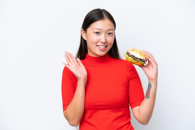Junge asiatische Frau, die einen Burger isoliert auf weißem Hintergrund hält und mit der Hand mit glücklichem Ausdruck salutiert