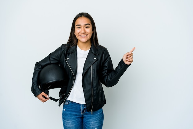 Junge asiatische Frau, die einen Motorradhelm über isolierte Wand hält, die lächelt und beiseite zeigt und etwas an der leeren Stelle zeigt