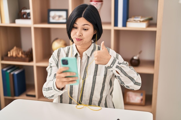 Junge asiatische frau, die einen videoanruf mit dem smartphone macht, das auf dem tisch sitzt und glücklich und positiv lächelt, den daumen hoch macht und ein zustimmungszeichen macht