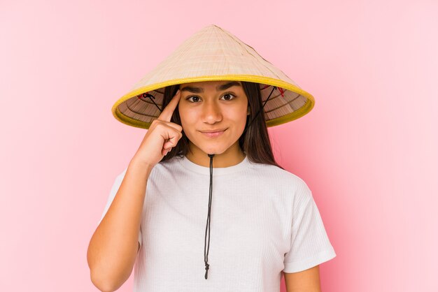 Junge asiatische Frau, die einen vietnamesischen Hut lokalisiert trägt Junge asiatische Frau, die einen vietnamesischen hatpointing Tempel mit dem Finger, denkend trägt, konzentrierte sich auf eine Aufgabe.