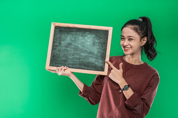 Junge asiatische Frau, die etwas auf einer leeren Tafel auf einer grünen Wand präsentiert und zeigt