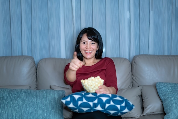 Junge asiatische Frau, die Fernsehspannungsfilm oder Nachrichten sieht, die glücklich und lustig schauen und Popcorn spät in der Nacht zu Hause Wohnzimmercouch während der Zeit der Hausisolation essen.