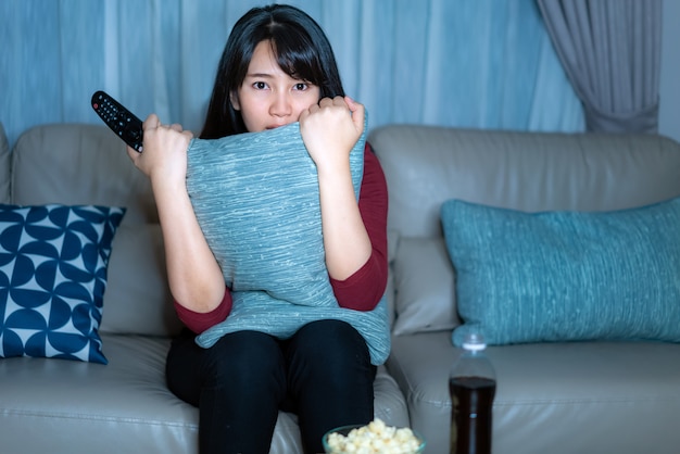 Junge asiatische Frau, die Fernsehspannungsfilm oder Nachrichten sieht, die schockiert und aufgeregt schauen, Popcorn spät in der Nacht zu Hause Wohnzimmercouch essend.