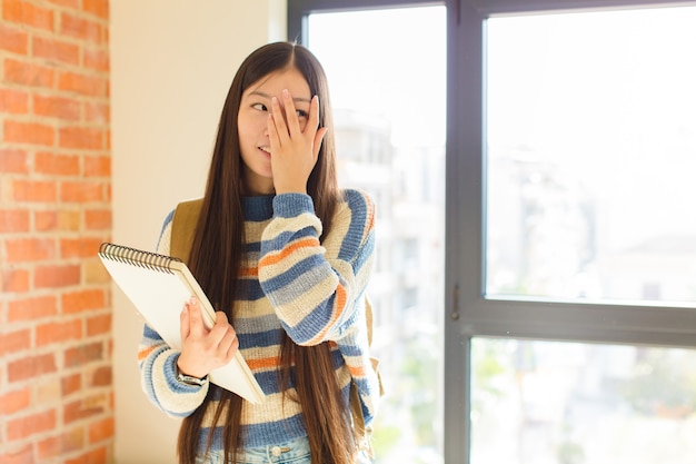 Junge asiatische Frau, die Gesicht mit Händen bedeckt, zwischen Fingern mit überraschtem Ausdruck späht und zur Seite schaut