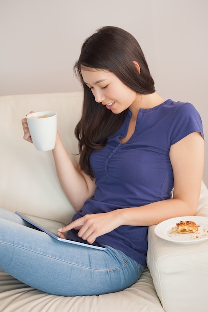 Junge asiatische Frau, die ihren Tabletten-PC verwendet und Becher Kaffee hält