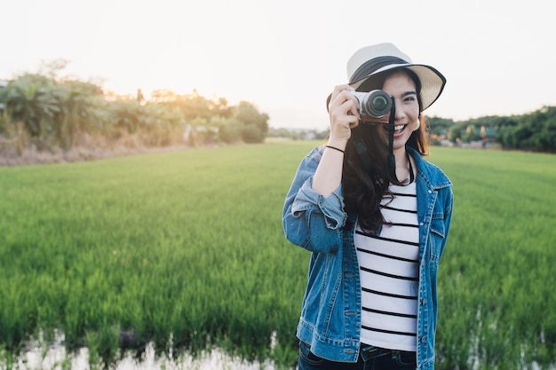 Junge asiatische Frau, die im Hut mit Kamera lächelt. Mädchen, das an der schönen Natur genießt