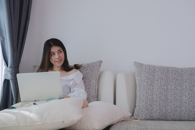 junge asiatische Frau, die Laptop auf Sofabett im Wohnzimmer denkt und verwendet.