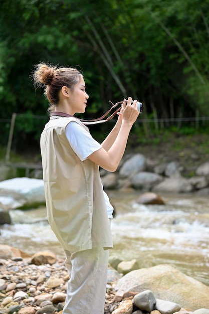 Junge asiatische Frau, die mit ihrer Retro-Filmkamera ein Foto des schönen Waldes und des Flusses macht
