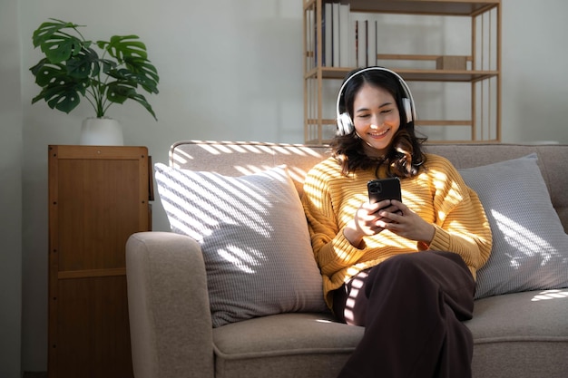 Junge asiatische Frau, die Musik über Kopfhörer hört und eine Notiz für ihre Arbeitsidee im Tagebuch schreibt, sitzt auf einem grauen Sofa im Wohnzimmer