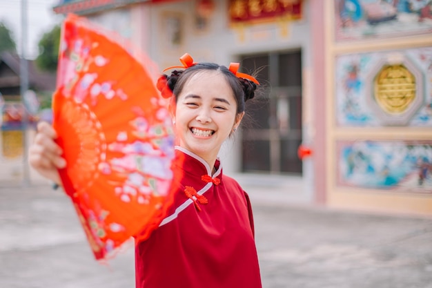 Junge asiatische Frau, die Qipao-Cheongsam-Kleid mit chinesischem Faltfächer auf rotem Hintergrund für Chine trägt