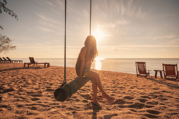 Junge asiatische Frau, die sich auf Holzschaukel am Strand bei Sonnenaufgang am tropischen Meer entspannt?