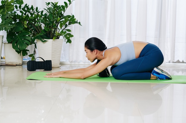 Junge asiatische Frau, die sich nach dem Training in Balasana oder Kindern entspannt, posieren auf Yogamatte zu Hause