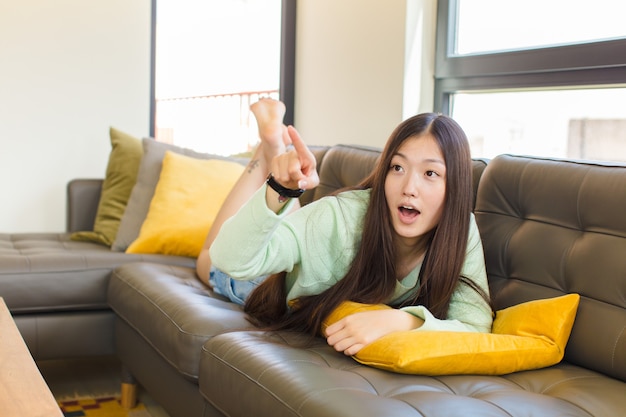 Junge asiatische Frau, die sich schockiert und überrascht fühlt, ehrfürchtig zeigt und mit erstauntem Blick mit offenem Mund nach oben schaut