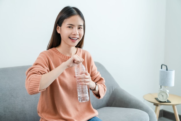 Junge asiatische Frau, die Wasserflasche und auf dem Sofa zu Hause hält.