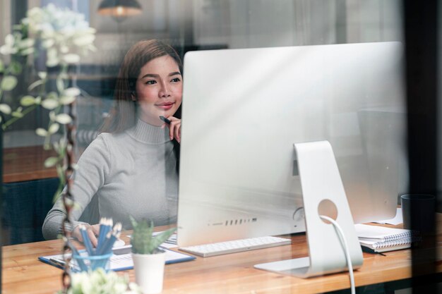 Junge asiatische Frau, die zu Hause am Desktop-Computer arbeitet, von zu Hause aus arbeitet, erfolgreiche Geschäftsfrau lächelt und Geschäftsergebnisse auf dem Bildschirm betrachtet.