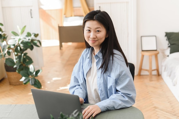 Junge asiatische Frau, die zu Hause an einem Laptop arbeitet und in die Kamera schaut Freiberufliches Geschäftskonzept