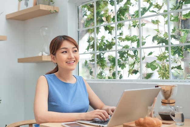 Junge asiatische Frau, die zu Hause arbeitet, während sie einen Laptop benutzt und hinter dem Fenster sitzt
