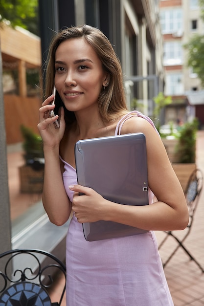 Junge asiatische Frau im Kleid, die im Freien steht, mit Laptop, der mit dem Smartphone anruft