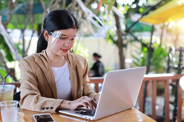 Junge asiatische Frau im lässigen Kleid mit Gesichtsschutz und Schutzmaske für das Gesundheitswesen, im Café sitzend und am Computer Laptop und Smartphone arbeitend. Neues normales und soziales Distanzierungskonzept