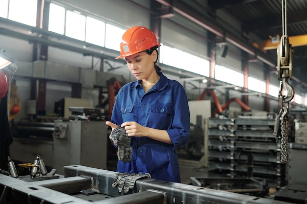 Junge asiatische Frau im orangefarbenen Helm, der Schutzhandschuhe beim Vorbereiten für Metallarbeiten im Fabrikladen trägt