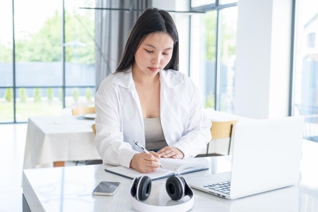 Junge asiatische Frau im weißen Hemd, die Notizen im Tagesbuch mit geöffnetem Laptop im modernen Zuhause macht