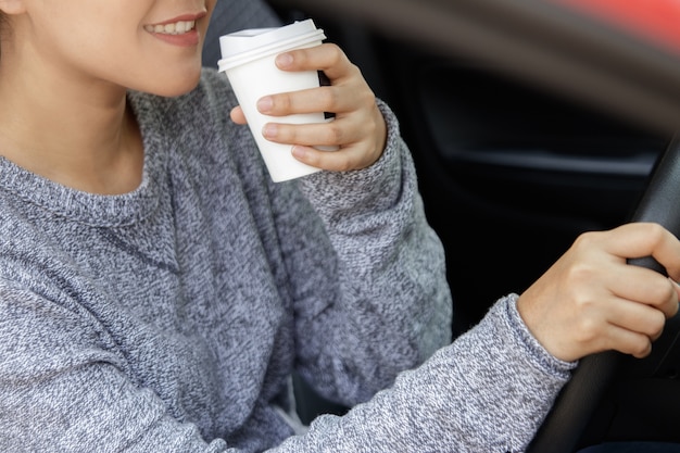 Junge asiatische Frau in der Freizeitkleidung, die ein Auto antreibt und Kaffee trinkt.