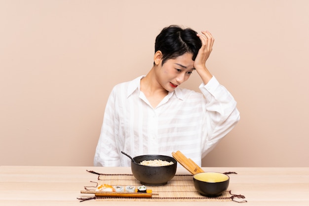 Junge asiatische Frau in einem Tisch mit Schüssel Nudeln und Sushi, die Zweifel mit verwirrendem Gesichtsausdruck haben