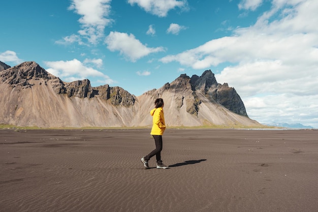 Junge asiatische Frau in gelber Jacke, die am Strand mit dem Berg Vestrahorn im Wikingerdorf auf Stokknes in Island steht